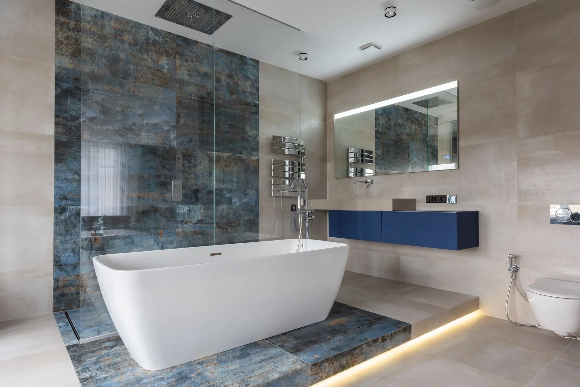 A white freestanding bath on a raised plinth tiled in blue-grey stone, with an LED-lit mirror and a navy floating vanity behind