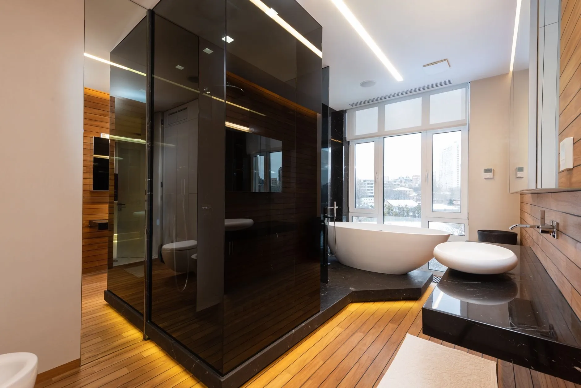 A bathroom with a smoked-glass shower enclosure on a lit timber plinth, a freestanding bath by tall windows and walnut-clad walls