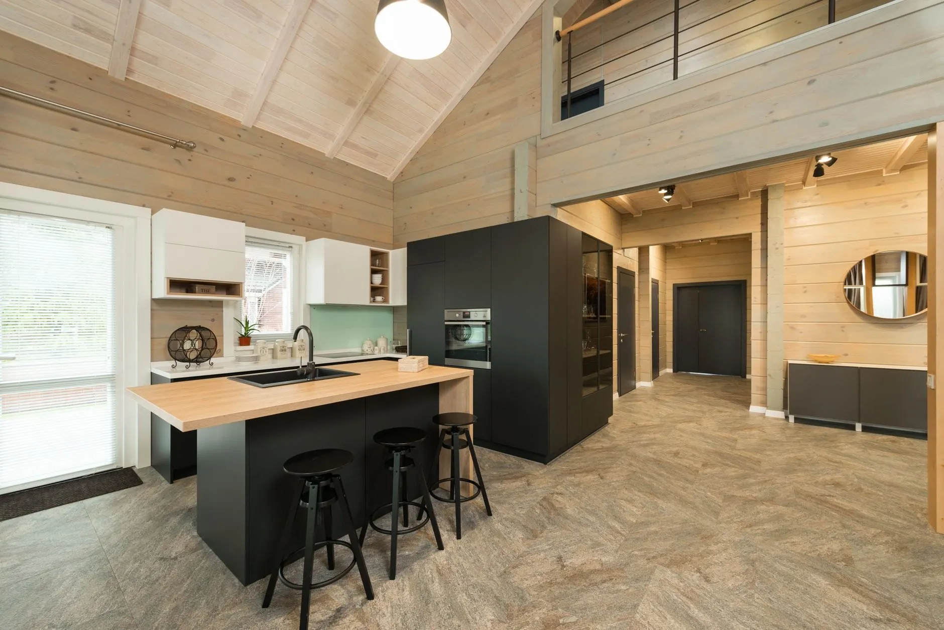 An open-plan kitchen under a vaulted timber ceiling, with a black island topped in pale oak, three black stools and a tall dark unit housing the oven