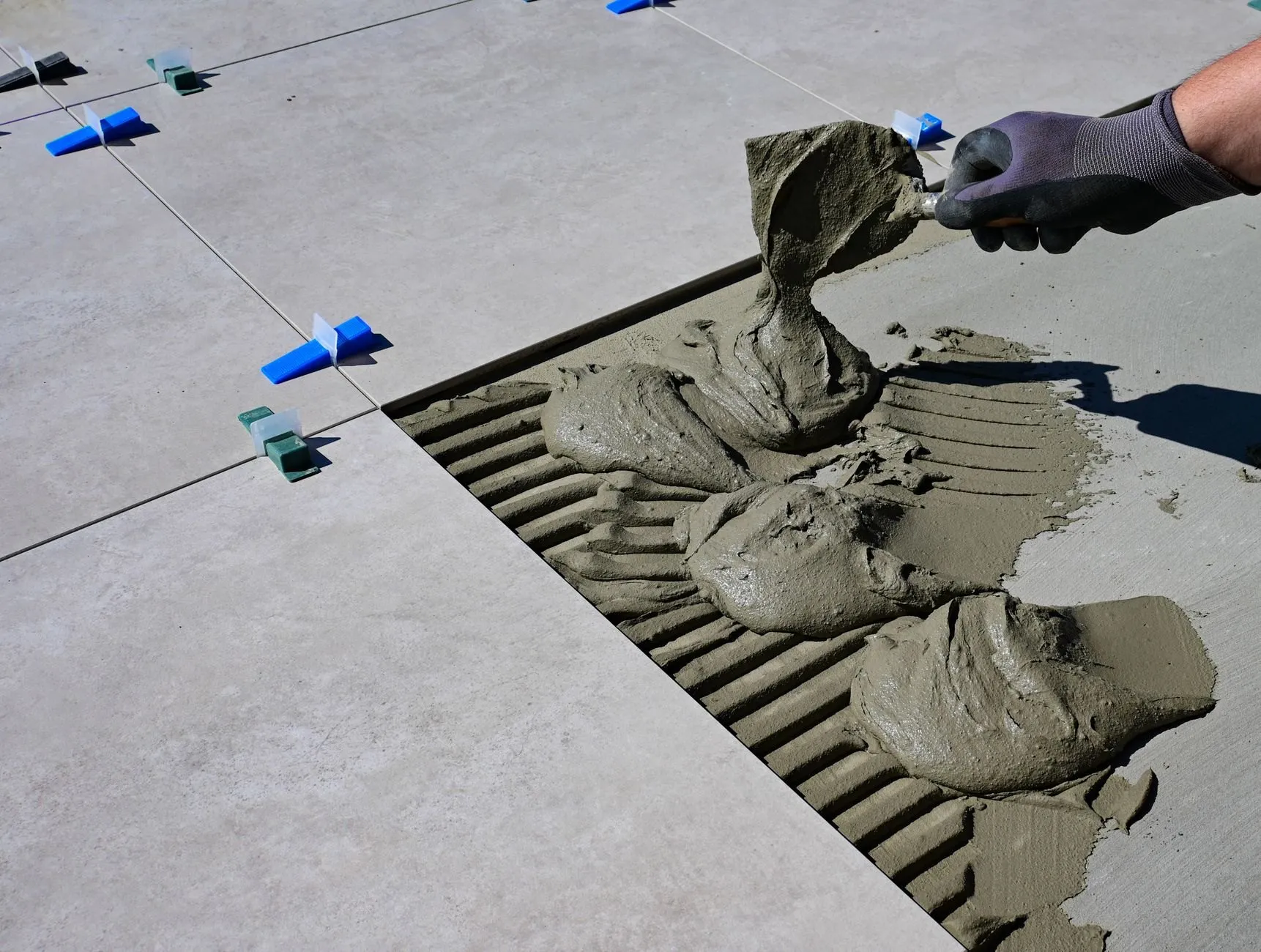 A tiler’s gloved hand spreading adhesive with a trowel between large grey porcelain floor tiles held level with clips